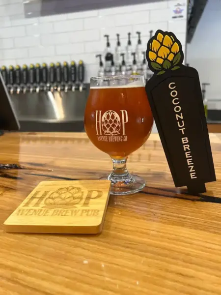 "Coconut Breeze fruited wheat beer in a branded glass next to its tap handle at Hop Avenue Brew Pub in Bridge City."