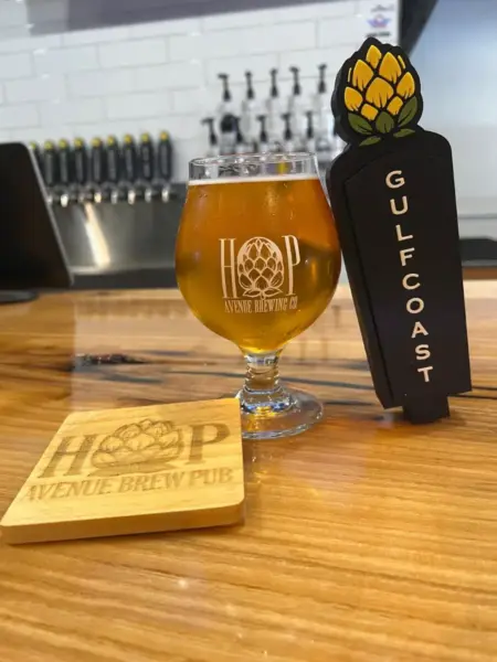 Gulf Coast Cream Ale in a branded glass next to its custom tap handle at Hop Avenue Brew Pub in Bridge City.