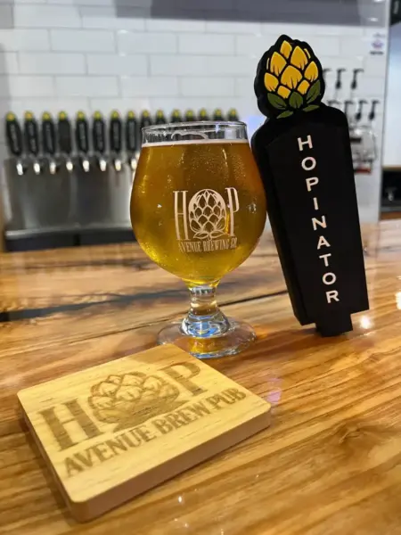 "Hopinator American IPA in a branded glass next to its custom tap handle at Hop Avenue Brew Pub in Bridge City."