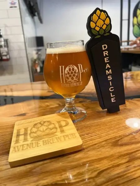 "Orangefield Dreamsicle Cream Ale in a branded glass next to its tap handle at Hop Avenue Brew Pub in Bridge City."