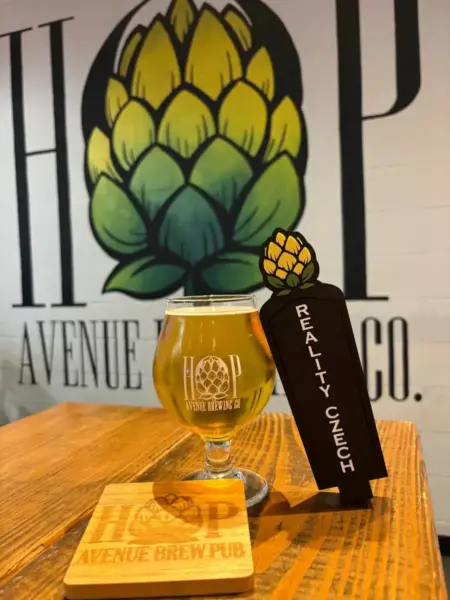 Reality Czech pilsner in a branded glass next to its tap handle at Hop Avenue Brew Pub in Bridge City