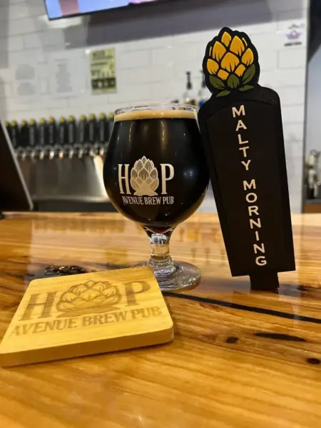 Malty Morning Dark Oatmeal Stout in a branded glass next to its tap handle at Hop Avenue Brew Pub in Bridge City.